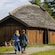 A man and a woman dressed in traditional Viking clothing, walking towards a traditional Viking farmhouse